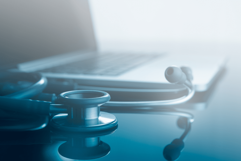 A stethoscope rests on a reflective surface in front of a blurred laptop, with a blue-tinted, hazy background.